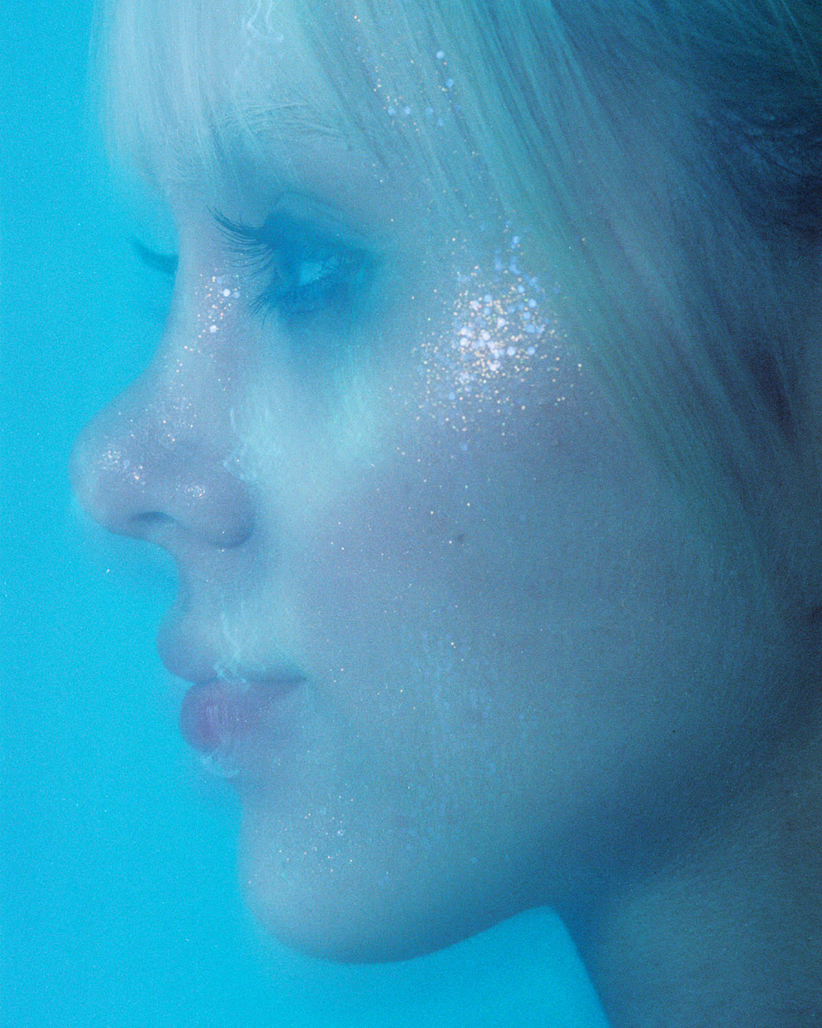 A close-up photo of a woman's face adorned with glitter against a blue backdrop. A combination of lens filters, post-processing, and shutter speed gives a washed-out and dreamy effect that almost gives the appearance of being taken underwater.